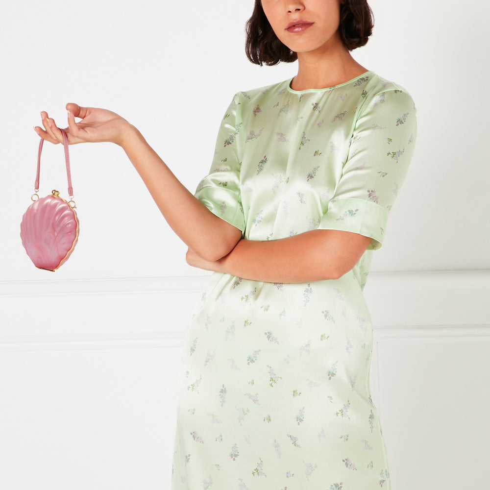 Woman in light green floral dress holding a pink seashell-shaped handbag.
