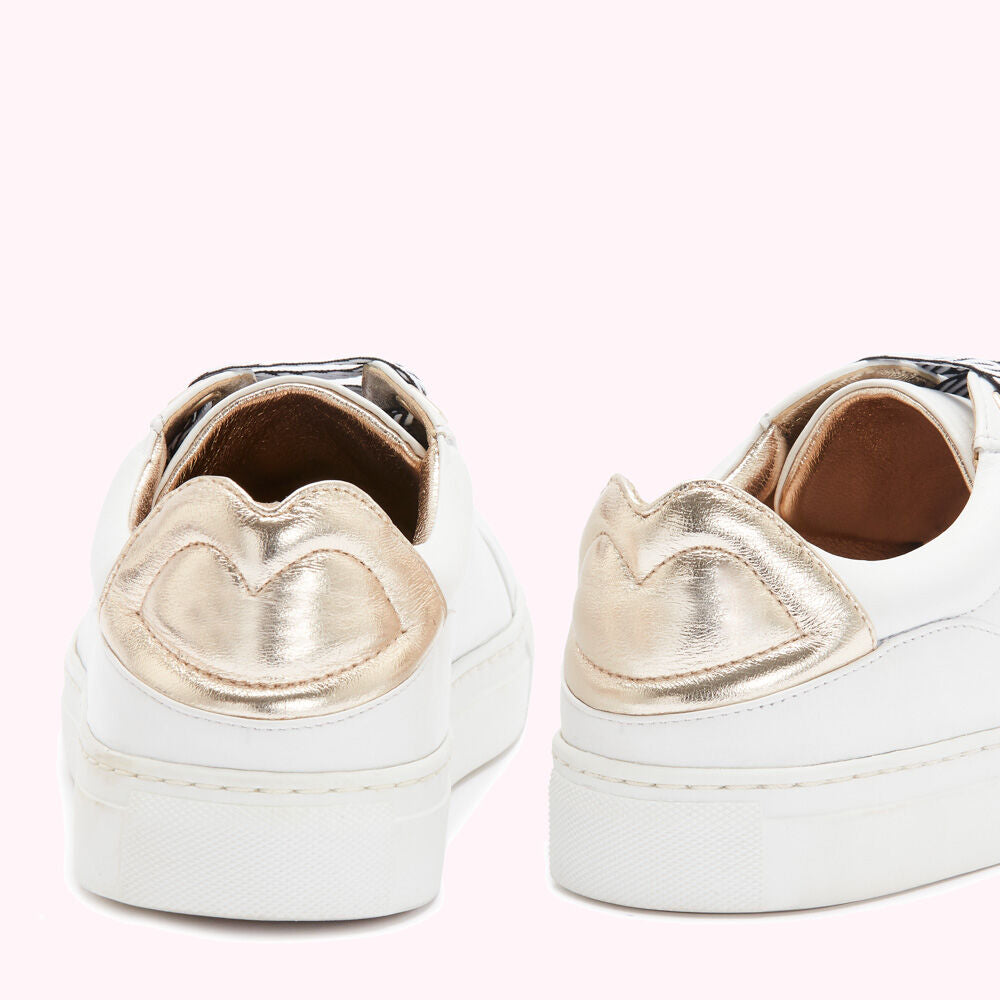 Rear view of a pair of white leather sneakers with rose gold heart-shaped accents on a pink background.

