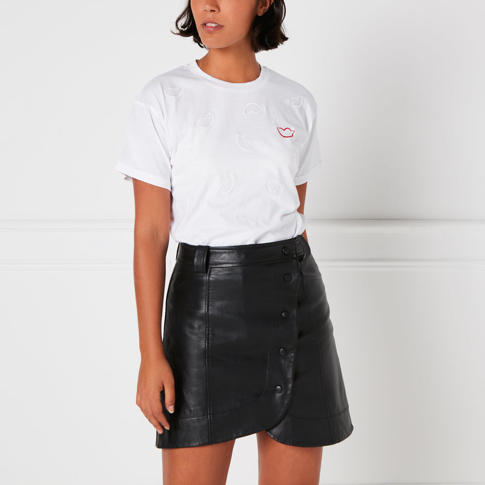Woman wearing a white t-shirt with embroidered lips and a black leather mini skirt against a white background.
