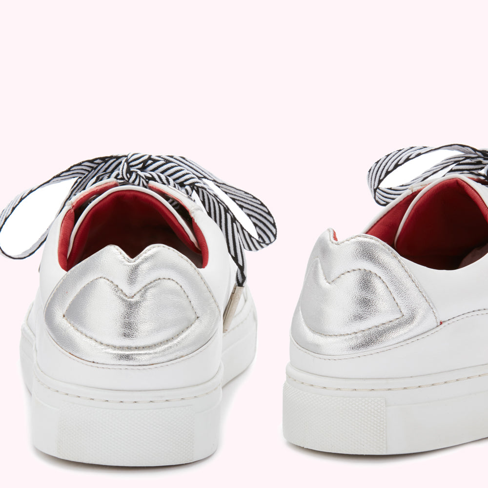 Pair of white sneakers with silver lip-shaped detail on the back and black and white striped laces against a pink background.
