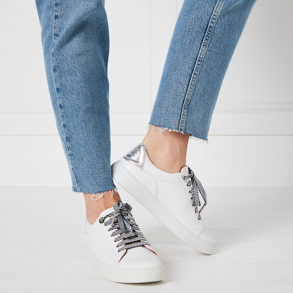 A pair of white leather sneakers with black and white striped laces, worn with light wash jeans against a white background.
