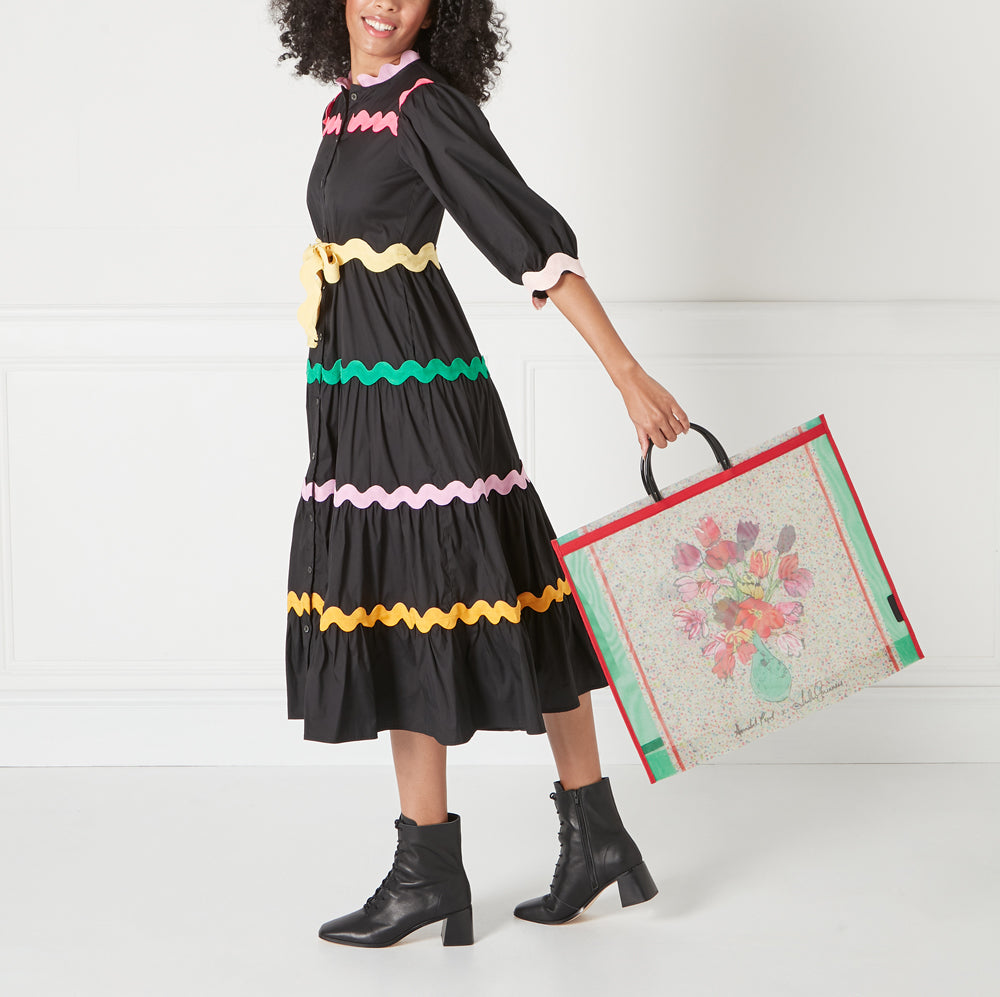 Woman in black dress with colorful trim holding a large tote bag with a floral print against a white background.
