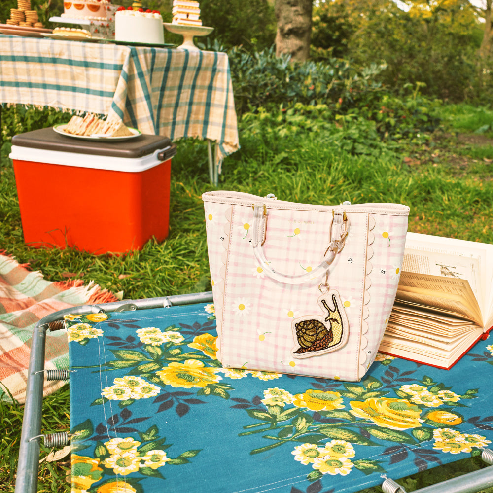 Pink gingham tote bag with snail charm on floral picnic blanket.
