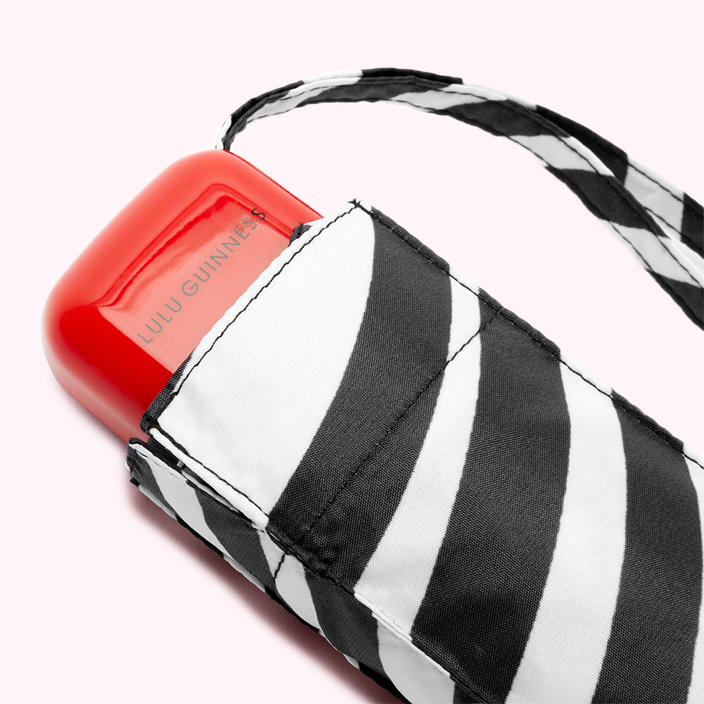 Close-up of a Lulu Guinness zebra-striped compact umbrella with its red case partially visible; the background is light pink.
