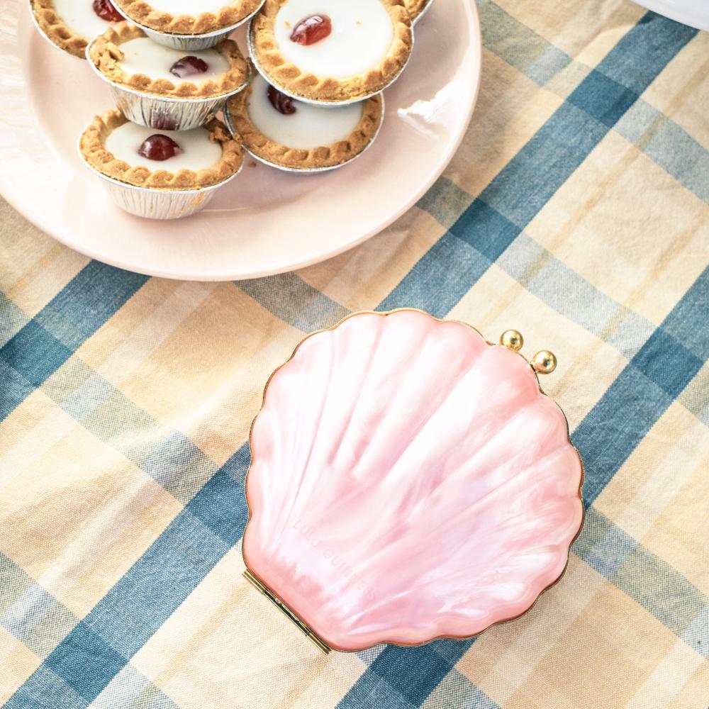 Pink seashell shaped box on a checkered tablecloth with a plate of mini tarts in the background. The shell has a gold hinge and two gold balls on the top.