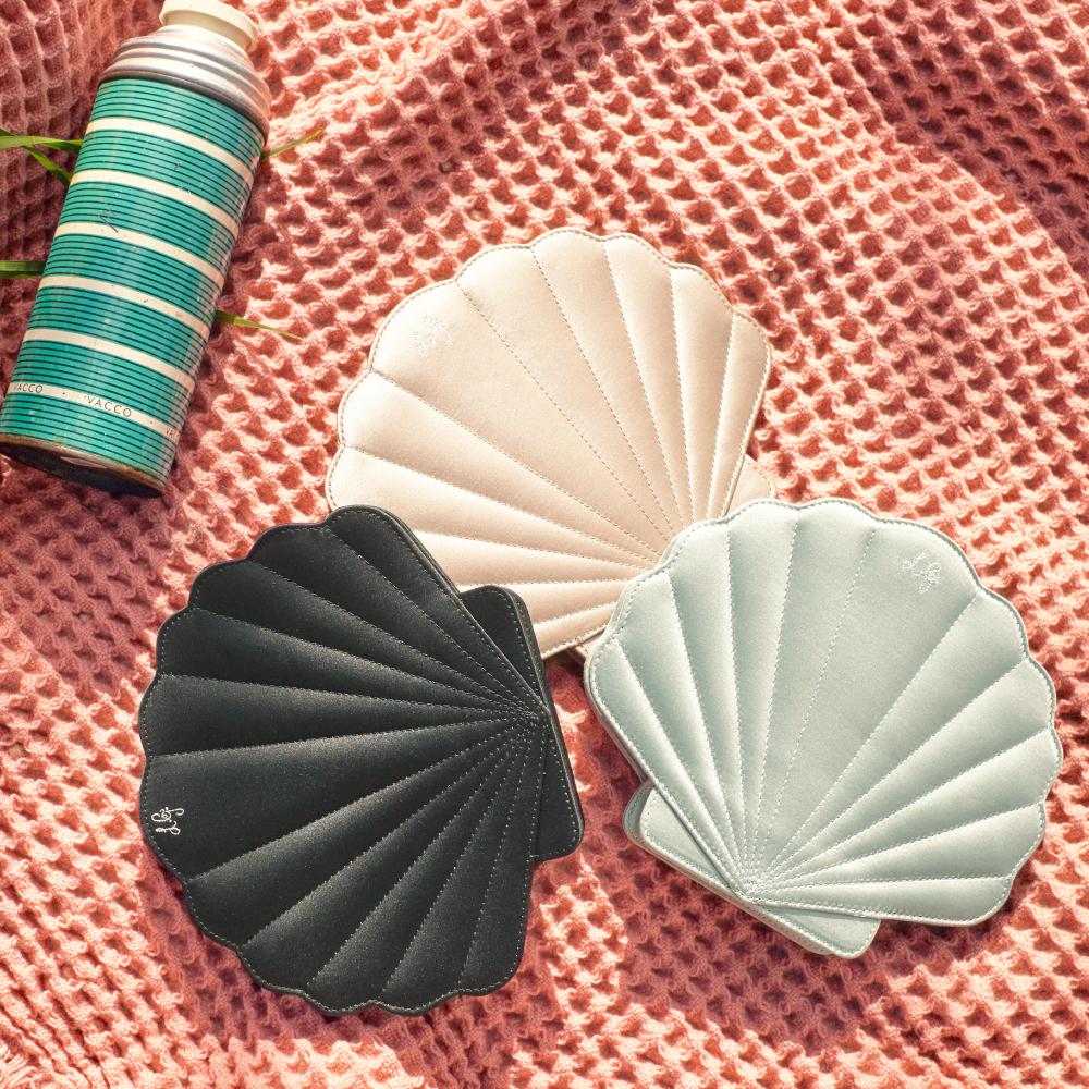 Three seashell-shaped compact mirrors in beige, black, and light green, arranged on a coral-coloured waffle knit fabric.