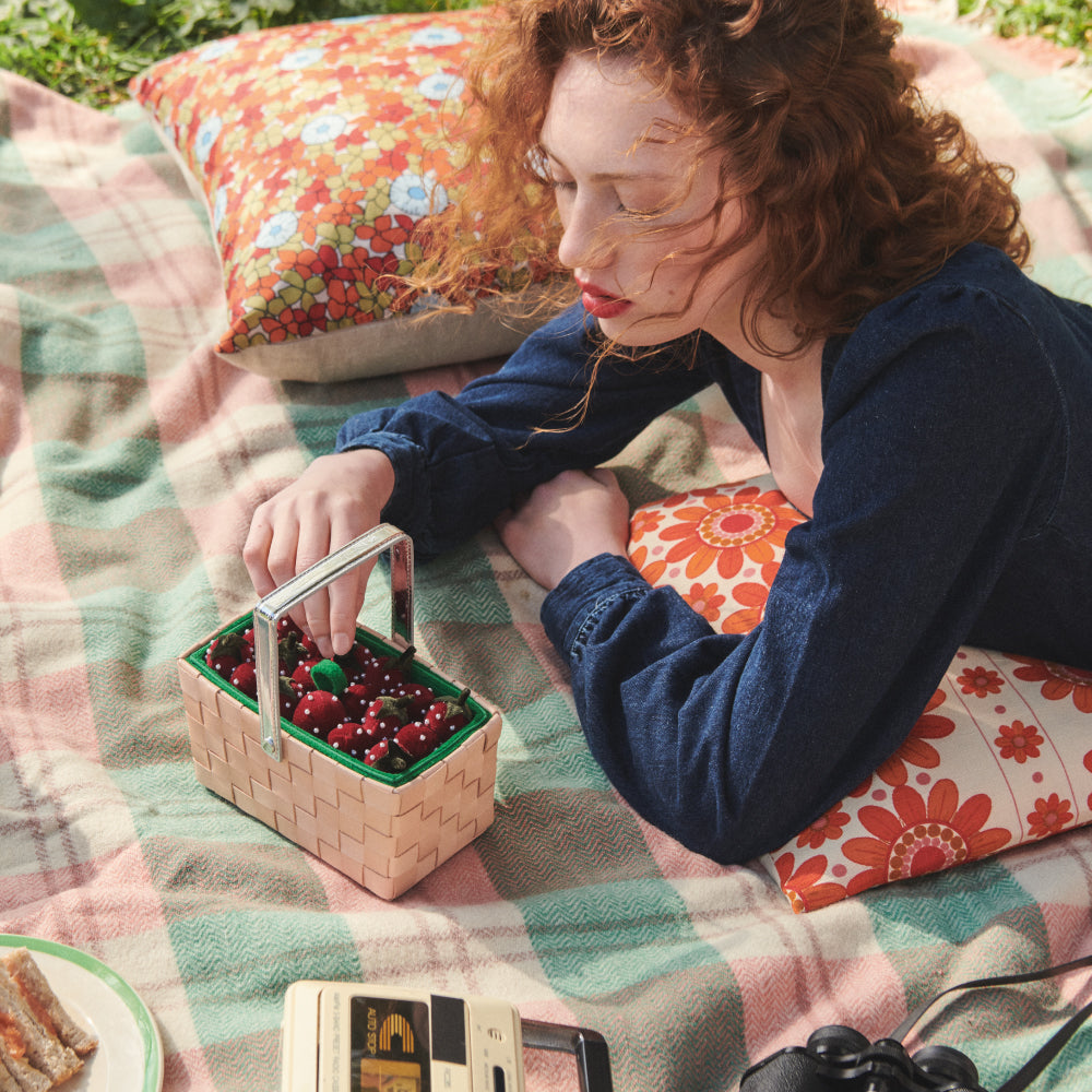 A red-haired woman lies on a picnic blanket, reaching into a small straw basket filled with strawberries.